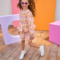 Child in a floral top and skirt wearing sunglasses posing in front of colorful geometric shapes.