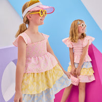Model in the front can be seen wearing woven tiered dress that features a stretchy ruching detail across bust and three tiers of a combination of pink/yellow/and blue floral patterns. Dress also features angel sleeve detail at straps for the finishing touch. Model in back can be seen wearing same floral print in our India top and tiered skort for a matching outfit moment.