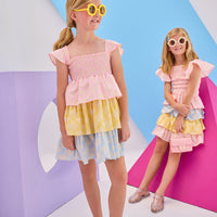 Model in the front can be seen wearing woven tiered dress that features a stretchy ruching detail across bust and three tiers of a combination of pink/yellow/and blue floral patterns. Dress also features angel sleeve detail at straps for the finishing touch. Model in back can be seen wearing same floral print in our India top and tiered skort for a matching outfit moment.