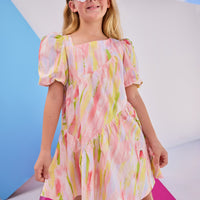 Model can be seen wearing woven dress in a bright pink/yellow/green watercolor print. Dress features a fun asymmetrical hem which adds a fun feature to the dress. Dress also has slight puff sleeves and also has a keyhole button closure on back of neckline.