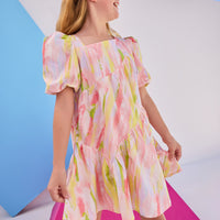 Model can be seen wearing woven dress in a bright pink/yellow/green watercolor print. Dress features a fun asymmetrical hem which adds a fun feature to the dress. Dress also has slight puff sleeves and also has a keyhole button closure on back of neckline.