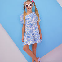 Model can be seen wearing a woven dress in a royal blue and white floral pattern. Dress features a fun asymmetrical hem which adds a fun feature to the dress. Dress also has slight puff sleeves and also has a keyhole button closure on back of neckline.