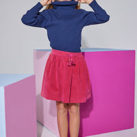 Tween skort in cranberry velvet which has fixed tie tassels in the front and an elastic waistband seen paired with a navy long sleeve turtleneck.