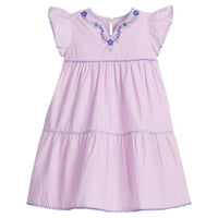 Girls/Tween woven purple tiered dress with hydrangea blue and purple embroidery detail on chest/along the v-neck neckline. Dress also features hydrangea pom poms along sleeve and first and bottom tiers of dress. A row of lilac pom poms is seen along the second tier of dress and around neckline. There is also a button closure at back of neckline for a secure fit.