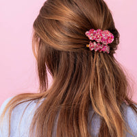 Woman with long brown hair styled in a half-up, half-down style with a pink floral hair accessory against a pink background.