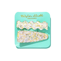 Two decorative hair clips with gold and white glitter on a teal card.