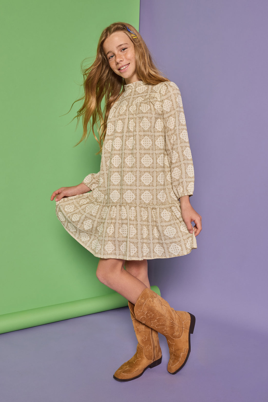 Model can be seen wearing a long sleeve woven dress in a light brown/cream print. Dress features high neck ruffle detail at collar and elastic sleeves for a relaxed fit.  
