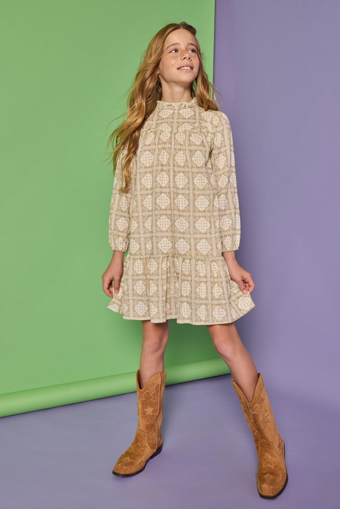 Model can be seen wearing a long sleeve woven dress in a light brown/cream print. Dress features high neck ruffle detail at collar and elastic sleeves for a relaxed fit.  