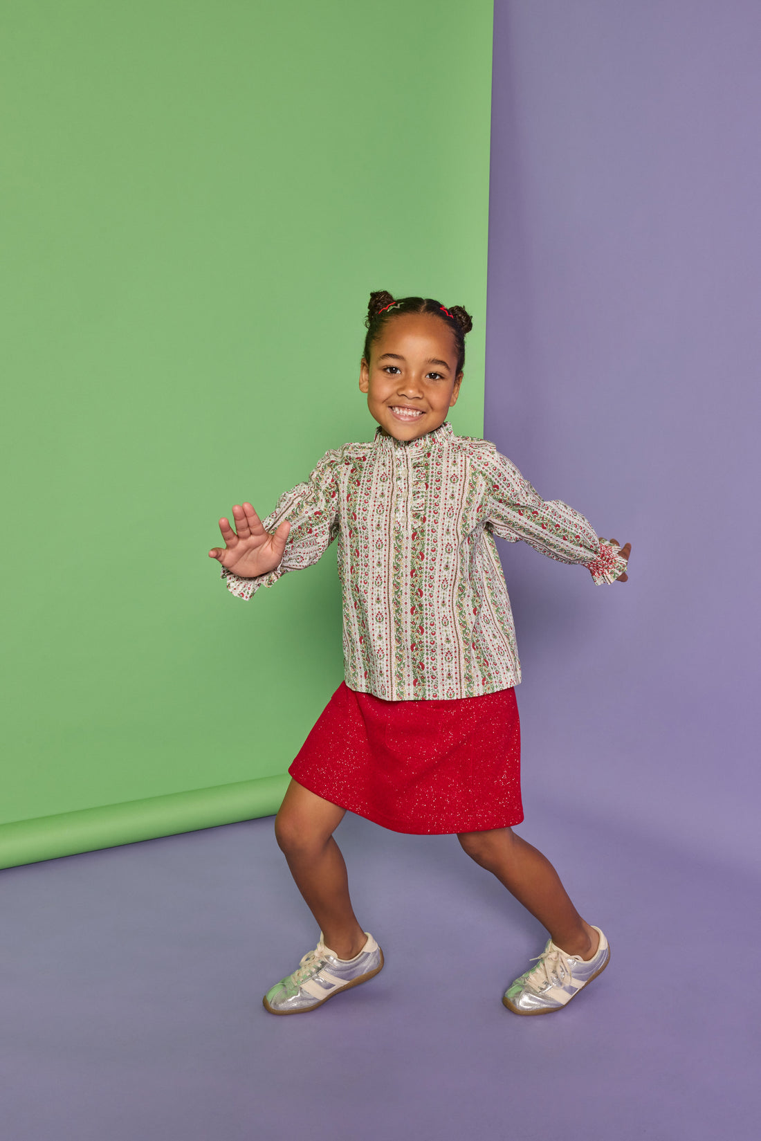 Model can be seen wearing a long sleeve green/red/white woven shirt that features button closures down the front, smocked red detail at sleeve cuffs, and ruffle detail at neckline. Paired back perfectly in our knit sparkly red skort with two pockets on the front to complete the look. 