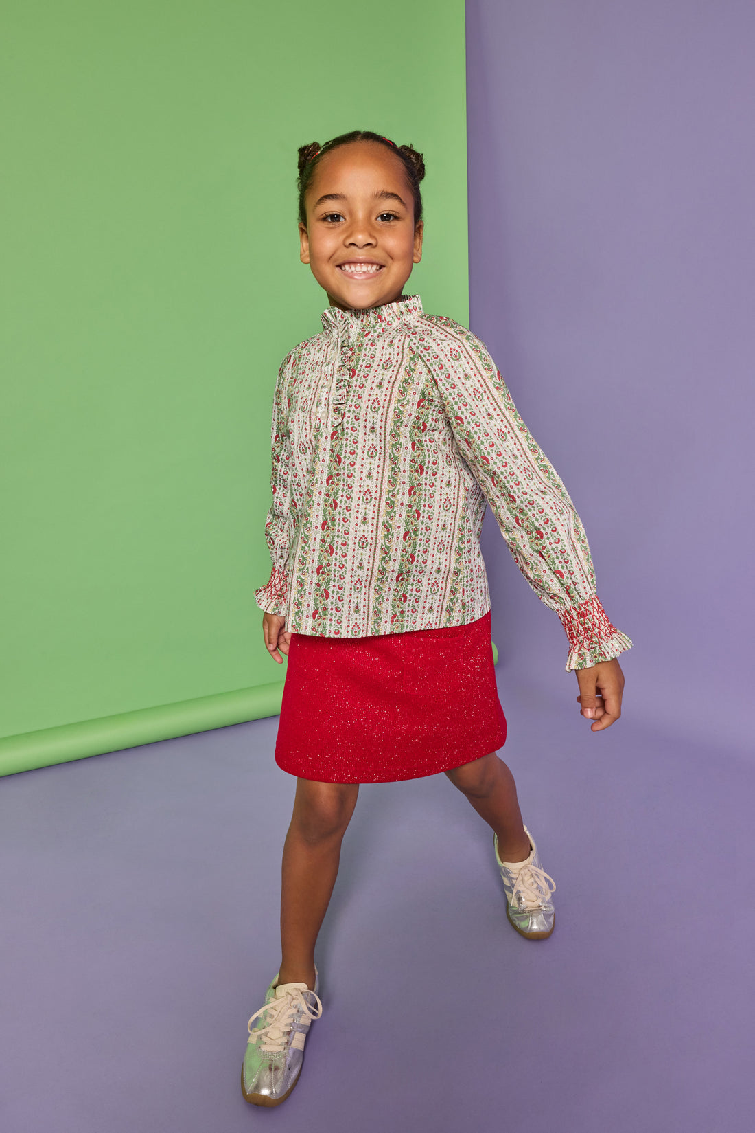 Model can be seen wearing a long sleeve green/red/white woven shirt that features button closures down the front, smocked red detail at sleeve cuffs, and ruffle detail at neckline. Paired back perfectly in our knit sparkly red skort with two pockets on the front to complete the look. 