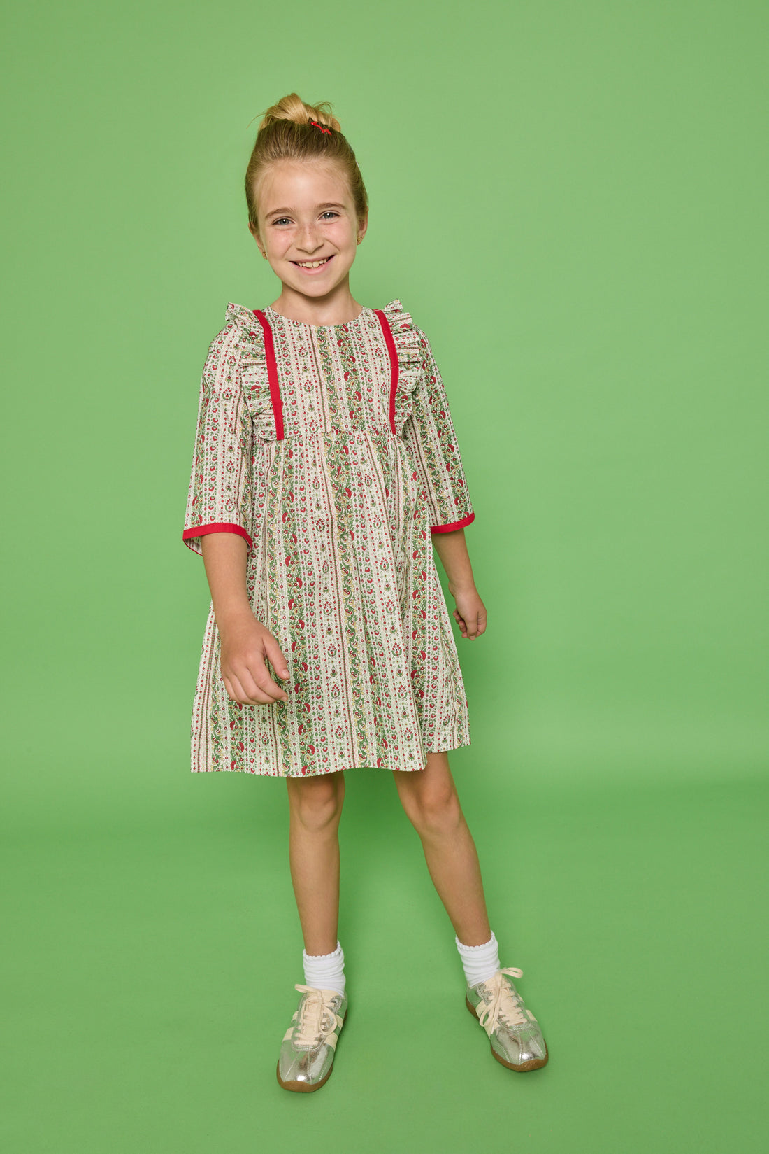 Model can be seen wearing a  3/4 sleeve woven dress in a green/red/white floral print. Dress features ruffle at top of sleeves and red ribbon detail at cuffs and side of bust for an elevated look. 