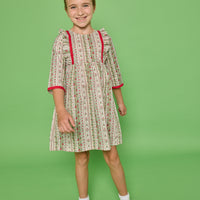 Model can be seen wearing a  3/4 sleeve woven dress in a green/red/white floral print. Dress features ruffle at top of sleeves and red ribbon detail at cuffs and side of bust for an elevated look. 