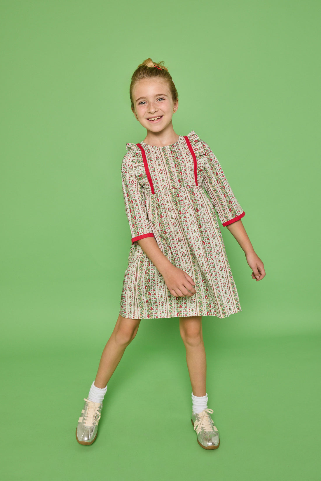 Model can be seen wearing a  3/4 sleeve woven dress in a green/red/white floral print. Dress features ruffle at top of sleeves and red ribbon detail at cuffs and side of bust for an elevated look. 