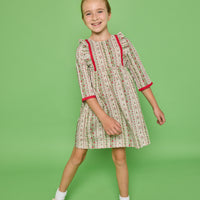 Model can be seen wearing a  3/4 sleeve woven dress in a green/red/white floral print. Dress features ruffle at top of sleeves and red ribbon detail at cuffs and side of bust for an elevated look. 