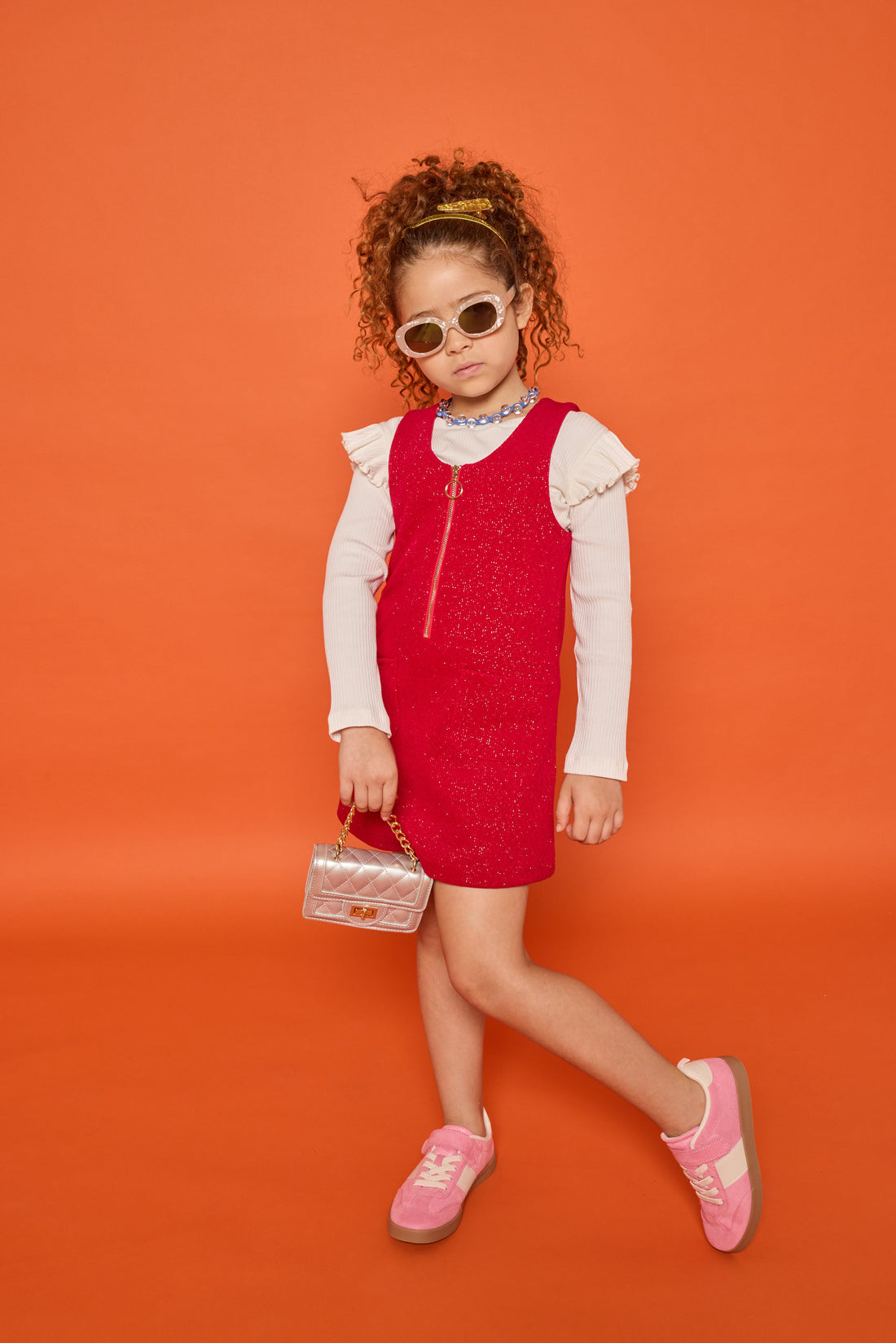 Model can be seen wearing white long sleeve ribbed knit top with ruffles at sleeves paired underneath cherry red sparkly knit jumper. Jumper features a quarter zip zipper and also features pockets at the front. 