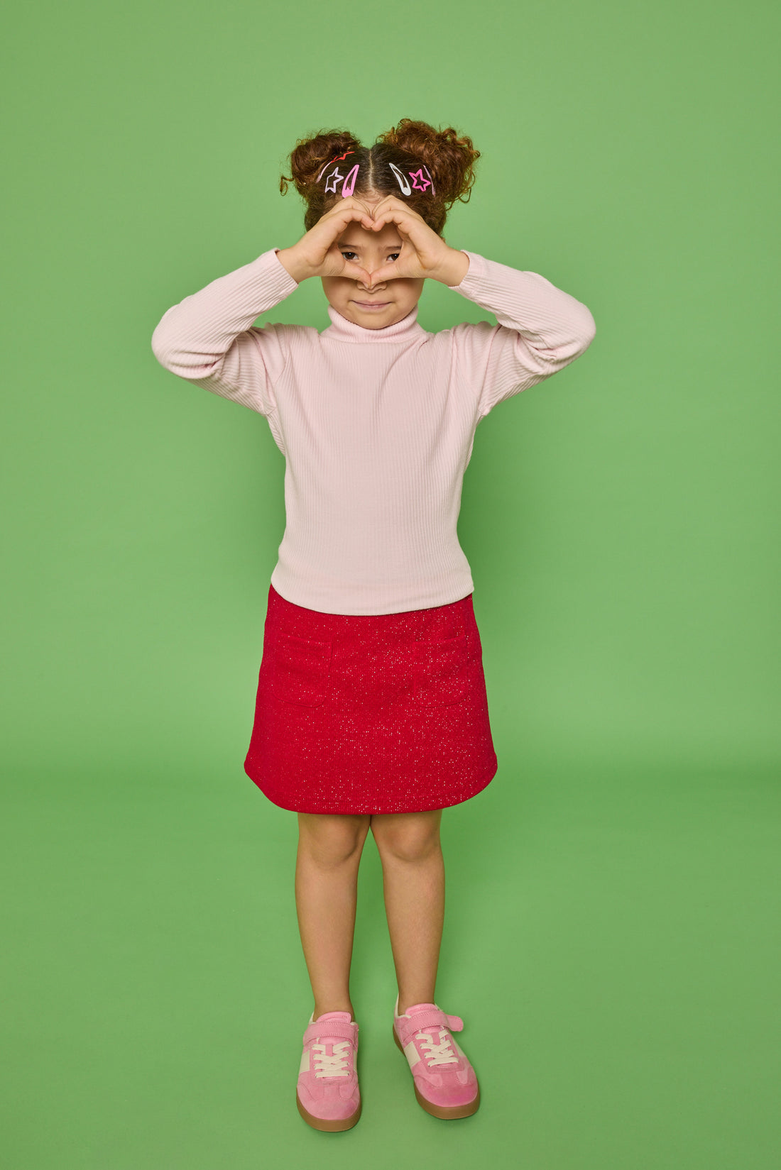 Model can be seen wearing a long sleeve ribbed knit turtleneck in bubblegum pink paired back with a red sparkly knit skort that has two pockets in the front and elastic waistband.
