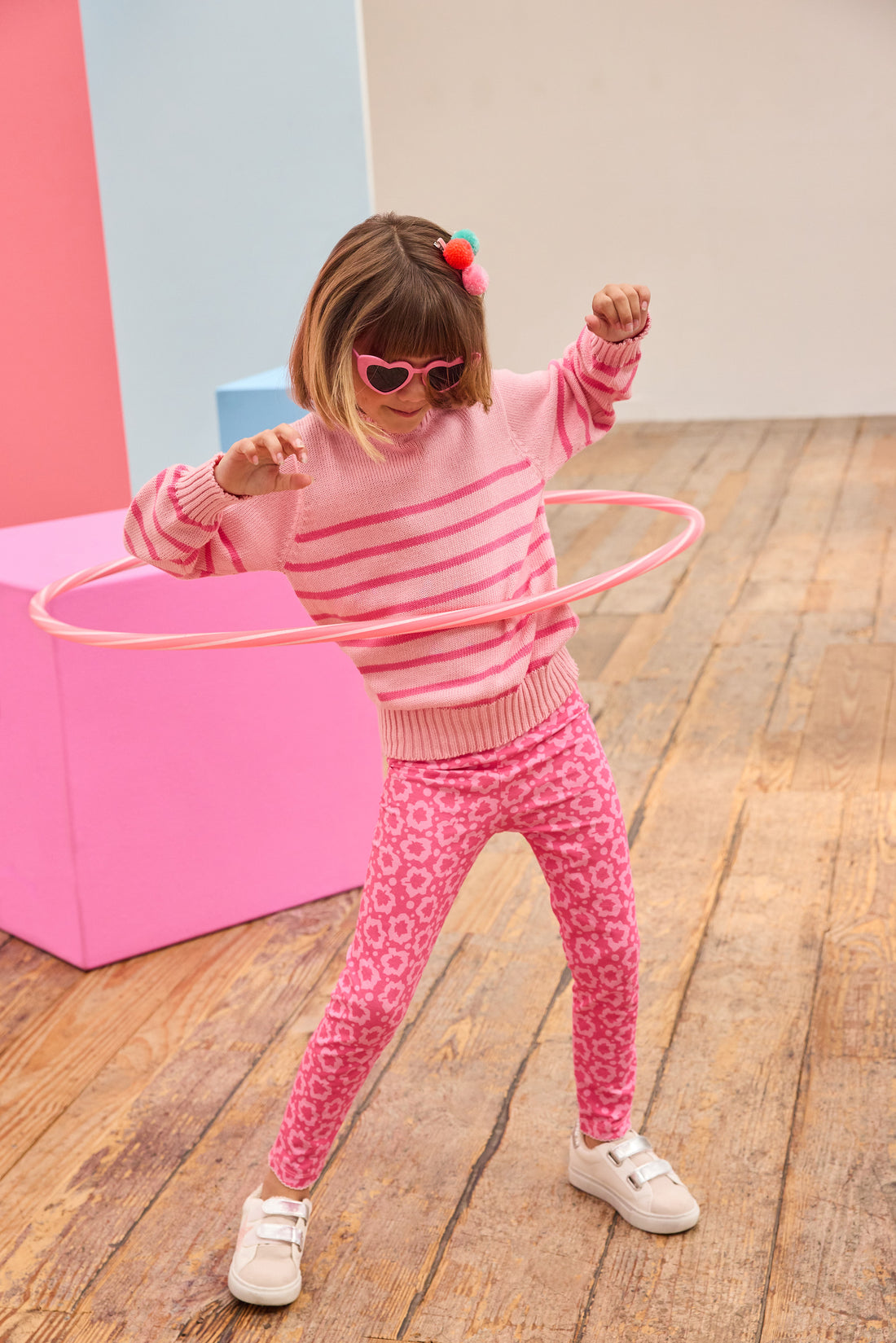 Child in pink striped sweater and floral print pants playing with a hula hoop on a wooden floor.