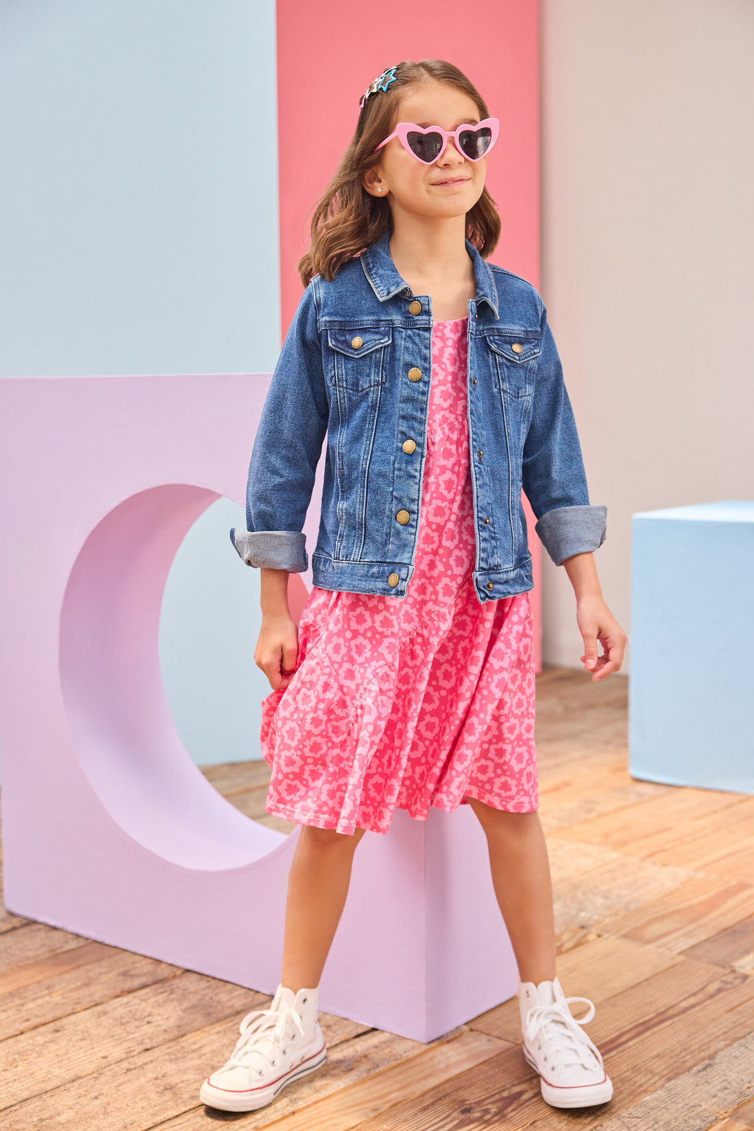 Young girl wearing a pink dress with a denim jacket and heart-shaped sunglasses in front of colorful geometric shapes.