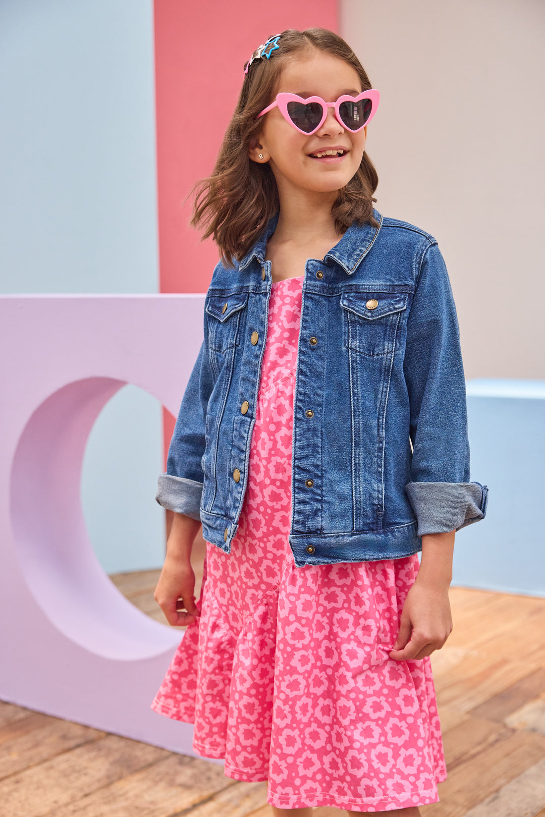 Young girl wearing a pink dress with a floral pattern and a blue denim jacket, standing against a colorful background.