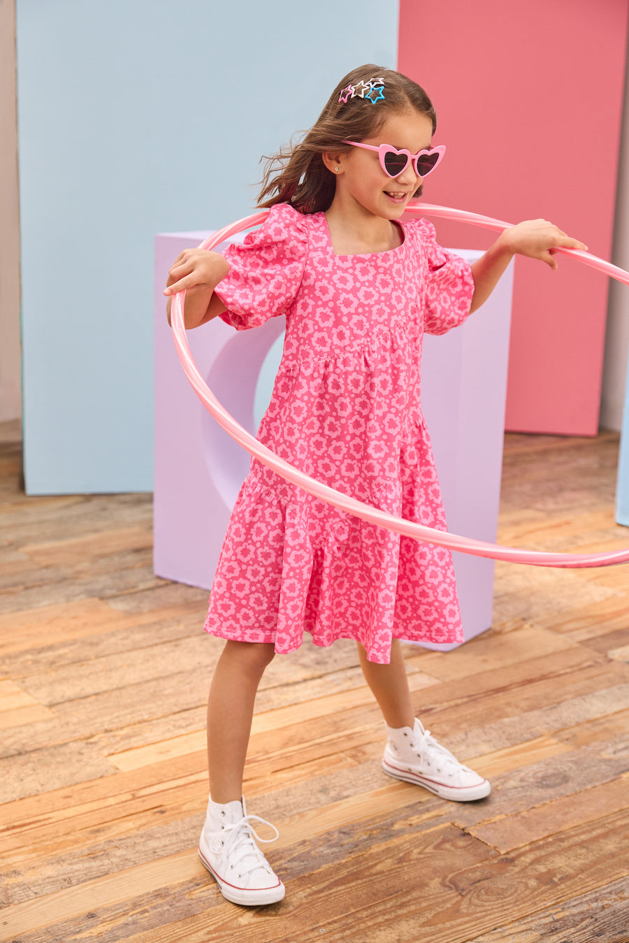 Young girl in a pink dress with a floral pattern, holding a pink hula hoop on a wooden floor.