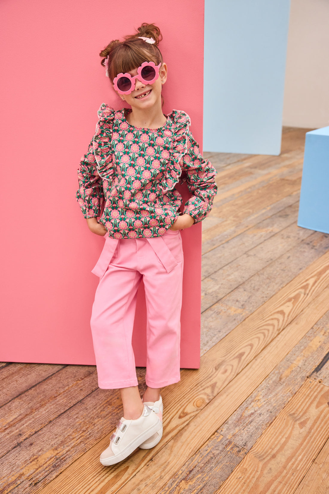 Model can be seen wearing pink/green/dark magenta floral woven top that has ruffle detail down the front of shirt for an elevated look, pairs back perfectly with bubblegum pink twill pants to complete the outfit. 