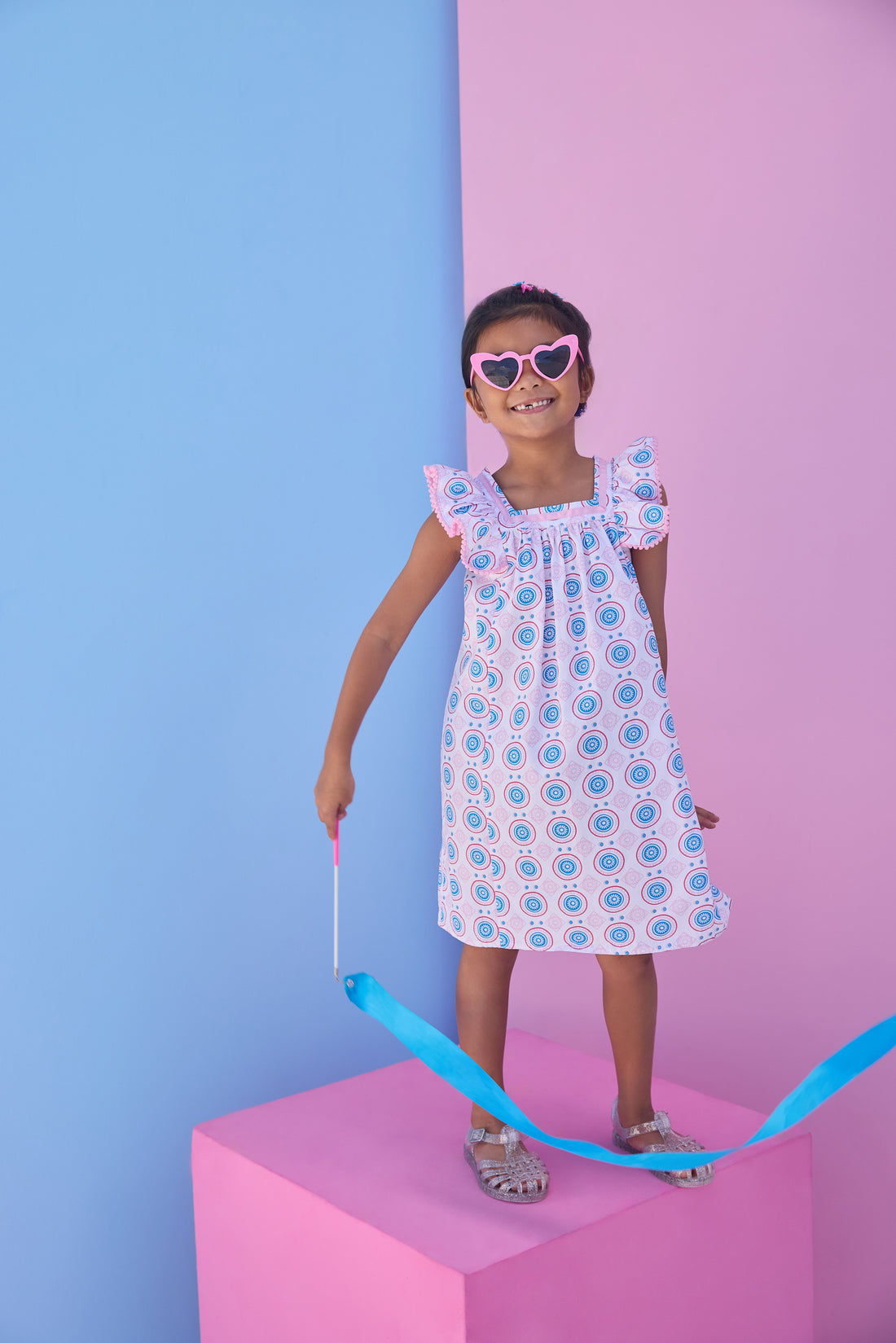 Model can be seen wearing a woven dress in a fun blue/pink/red print. Dress features a square neckline with pink ribbon detail across edge, pink pom pom detail at end of sleeves, and and angel sleeve detail to elevate the look. 