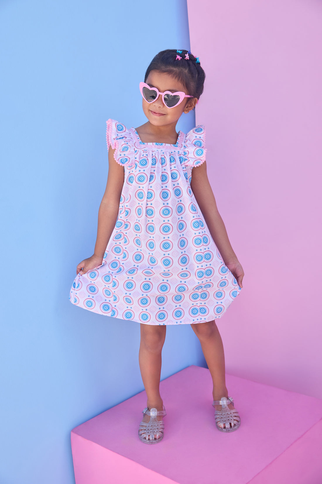 Model can be seen wearing a woven dress in a fun blue/pink/red print. Dress features a square neckline with pink ribbon detail across edge, pink pom pom detail at end of sleeves, and and angel sleeve detail to elevate the look. 