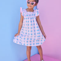 Model can be seen wearing a woven dress in a fun blue/pink/red print. Dress features a square neckline with pink ribbon detail across edge, pink pom pom detail at end of sleeves, and and angel sleeve detail to elevate the look. 