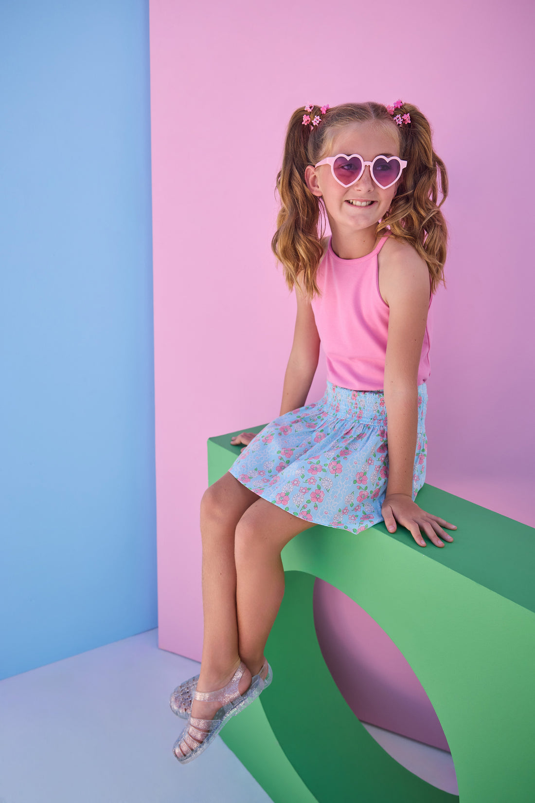 Tween model can be seen wearing a smocked waist woven skirt in a pink and blue floral print. Skirt features smocking detail at waist for a secure but comfy fit. Skirt pairs back perfectly with a hot pink halter top that has button closures along back. 