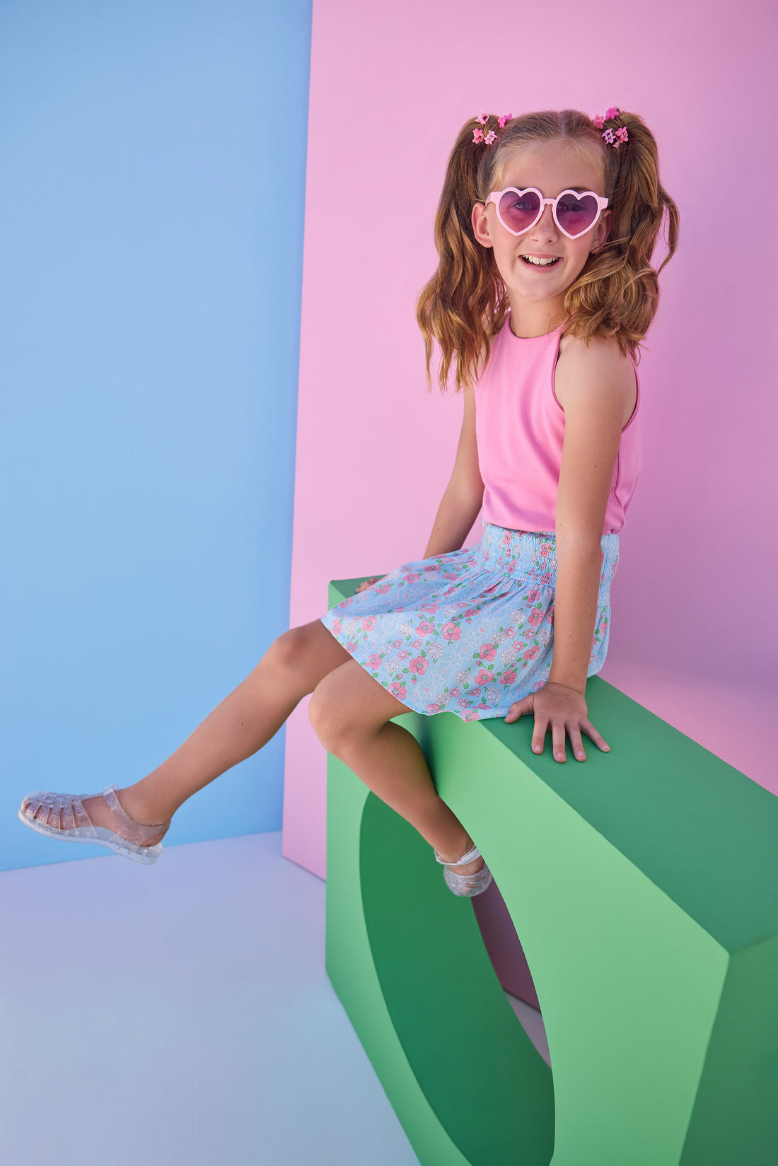 Tween model can be seen wearing a smocked waist woven skirt in a pink and blue floral print. Skirt features smocking detail at waist for a secure but comfy fit. Skirt pairs back perfectly with a hot pink halter top that has button closures along back. 