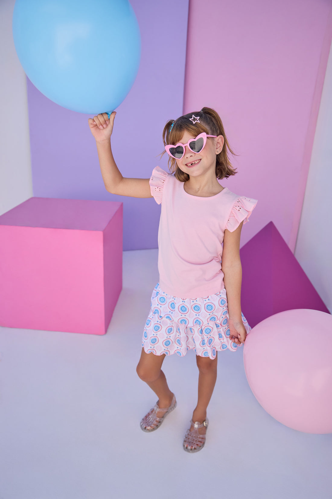 Model can be seen wearing two tiered skort in a blue/red/pink pattern that features scallop edges in pink embroidery, built in shorts, and an elastic waistband. Skort pairs back perfectly with the knit light pink top that features an eyelet detail on the angel sleeves to complete the look. 