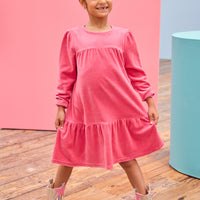 - [ ] Model can be seen wearing long sleeve velour dress in periwinkle blue. Dress features button closure at back, and pleating detail across bust and bottom tier of dress for an elevated look. 