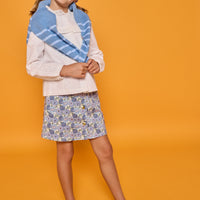 Model can be seen wearing an embroidered white woven top paired back with a corduroy skirt in a blue floral print. Model also has a sweater over her shoulders for an elevated look. 