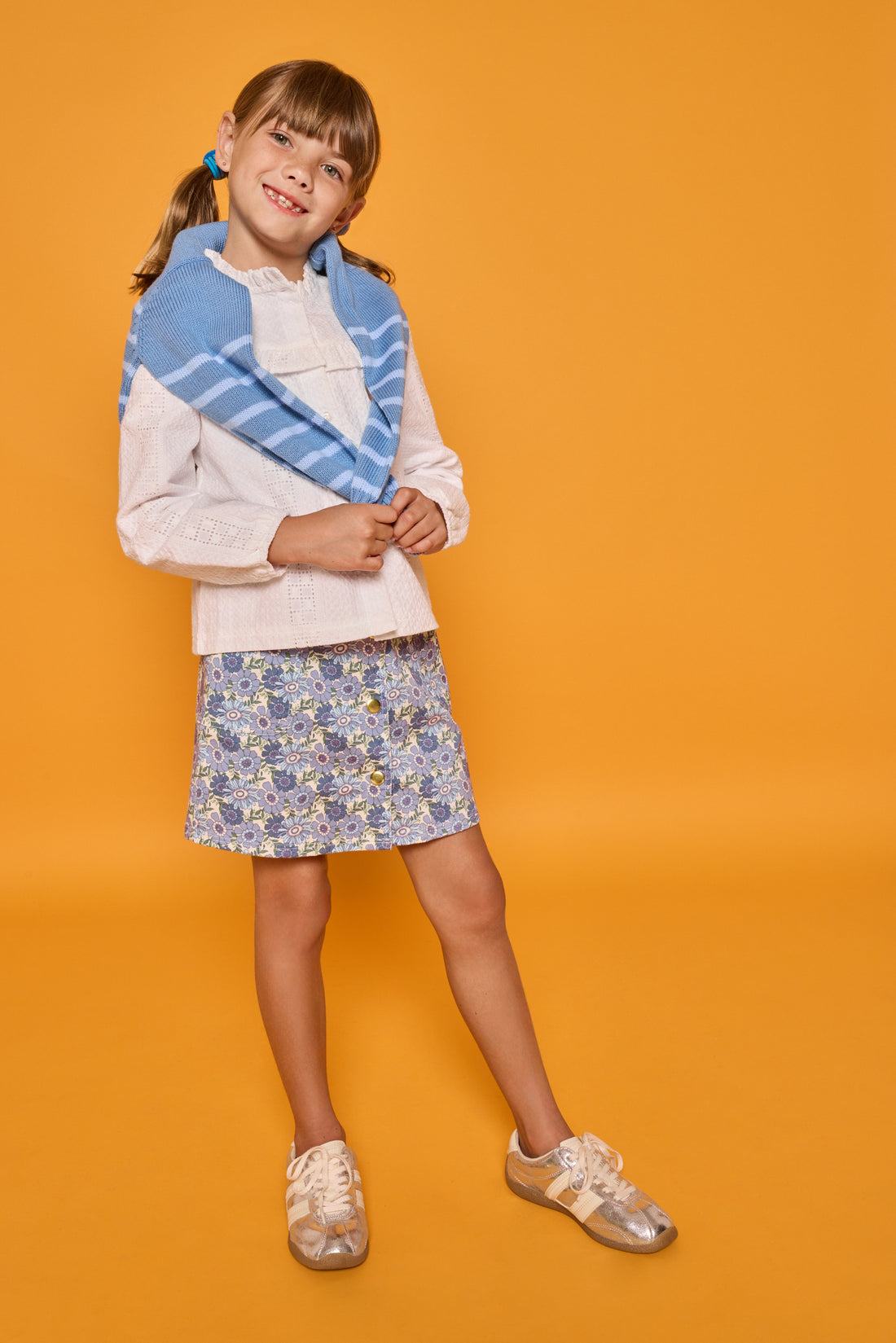 Model can be seen wearing white embroidered woven top with buttons down the front paired back with our corduroy skirt in a blue floral print. Also thrown over the shoulders is a knit striped sweater in blue and light blue stripe for an elevated look. 