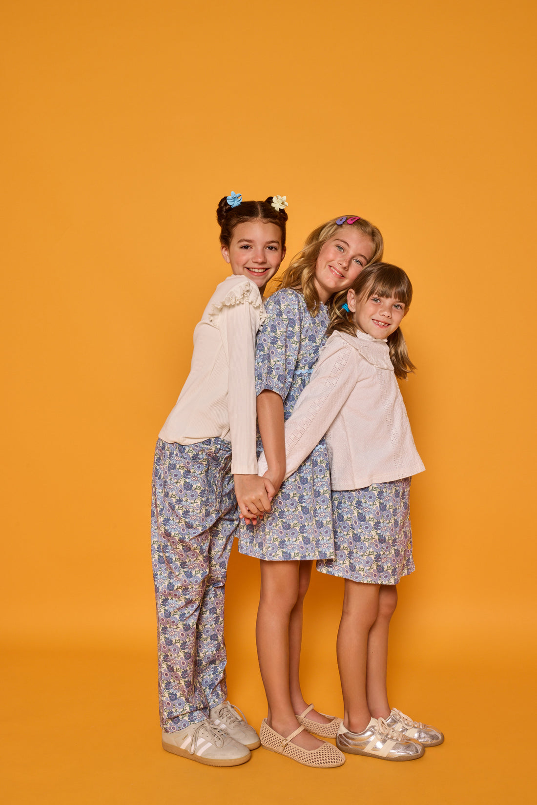 Model on left can be seen wearing white ribbed knit long sleeve top with ruffles at sleeves paired back with blue floral corduroy pants,  model in the middle can be seen wearing corduroy dress with 3/4 sleeves in the same floral print, and model on right can be seen in woven embroidered top in white paired back with same blue floral corduroy print skirt. 