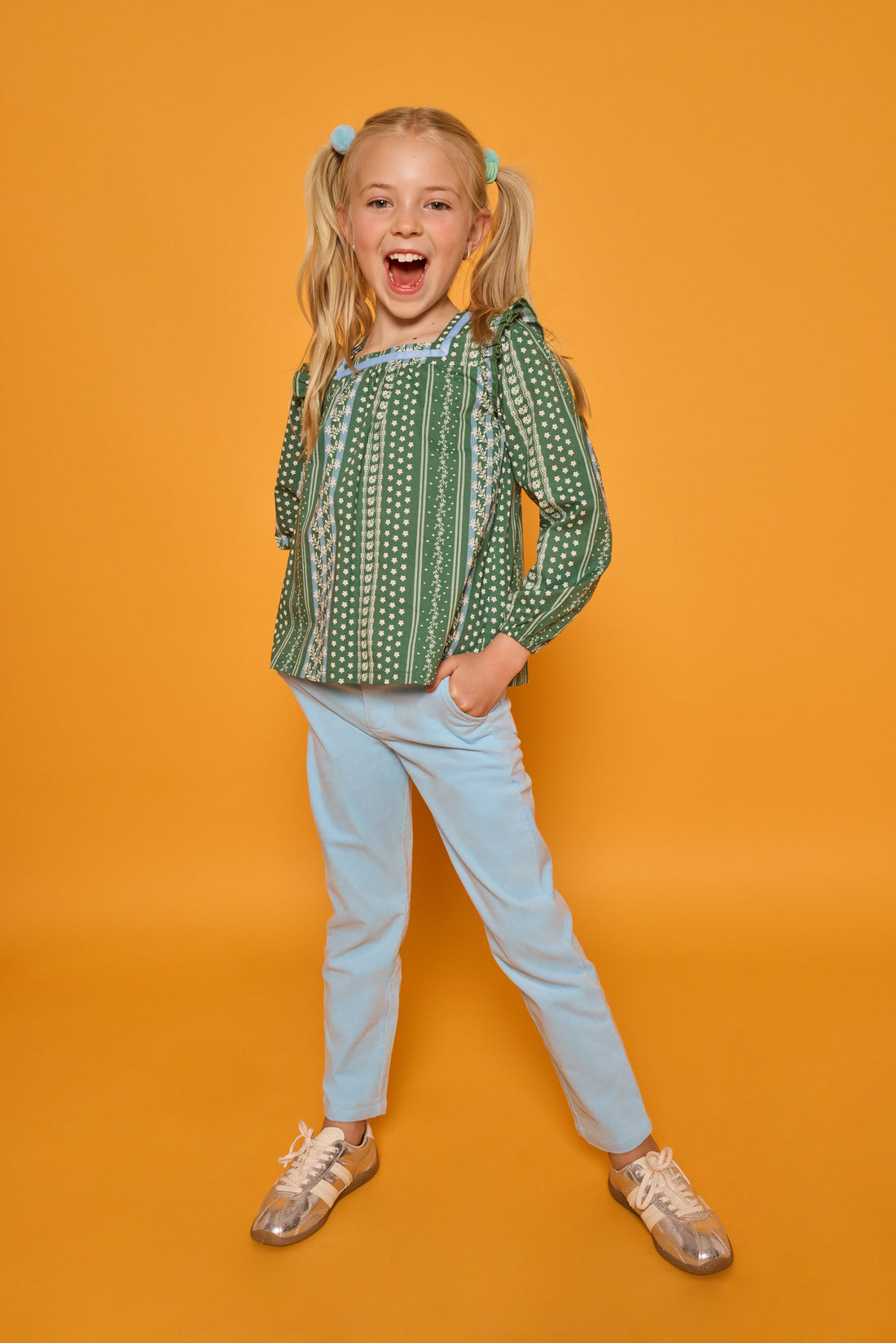 A young girl with blonde pigtails stands against a solid orange background, wearing a green and white patterned long-sleeve top, light blue pants, and metallic gold sneakers.