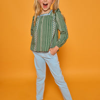 A young girl with blonde pigtails stands against a solid orange background, wearing a green and white patterned long-sleeve top, light blue pants, and metallic gold sneakers.