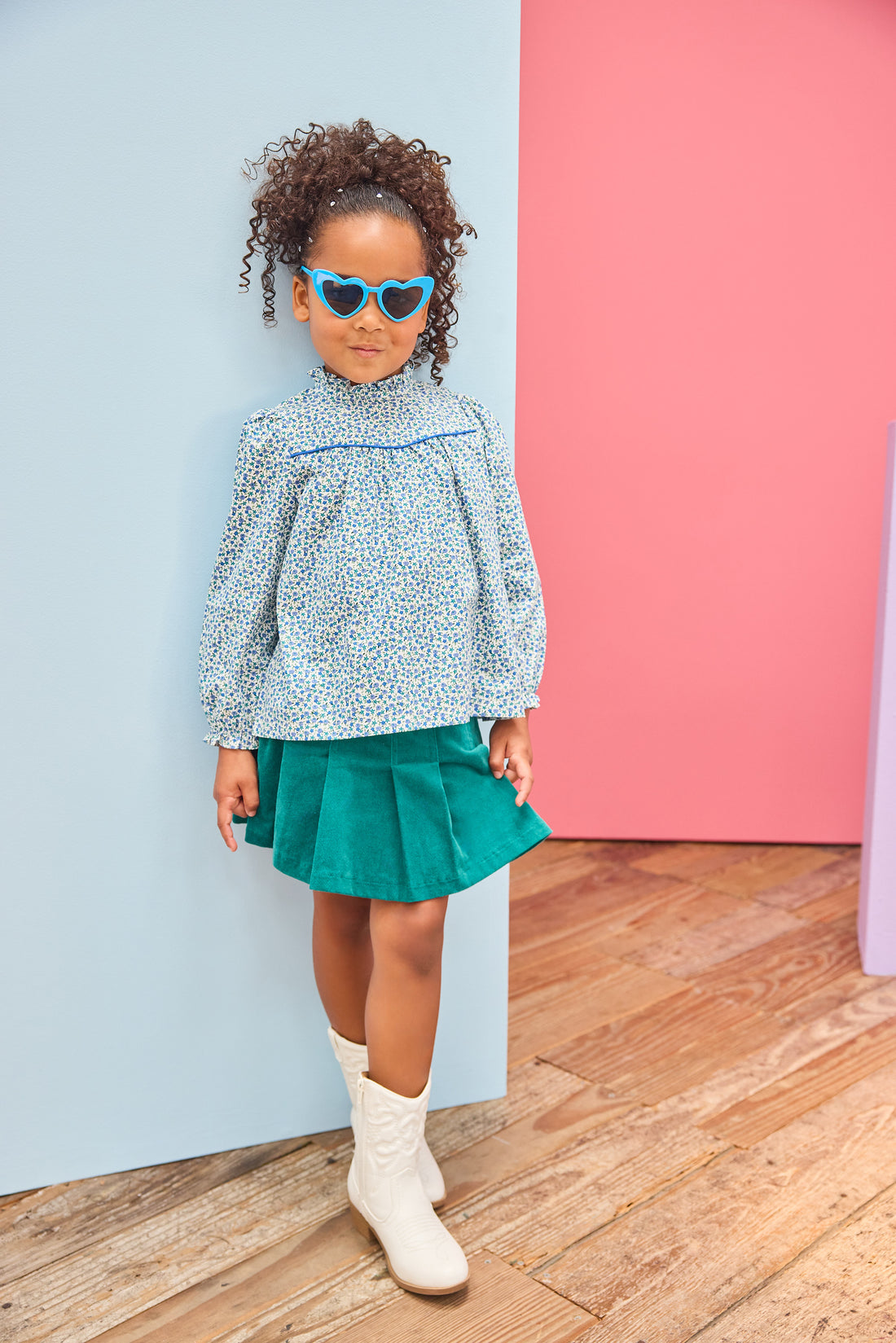 Model can be seen wearing a woven long sleeve top in a blue floral pattern that features a ruffle high neckline and blue trim/pleating detail and button closure along back. Paired back perfectly with a pleated skort in a green velvet to complete the look. 