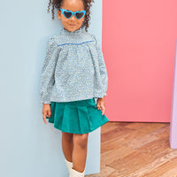 Model can be seen wearing a woven long sleeve top in a blue floral pattern that features a ruffle high neckline and blue trim/pleating detail and button closure along back. Paired back perfectly with a pleated skort in a green velvet to complete the look. 