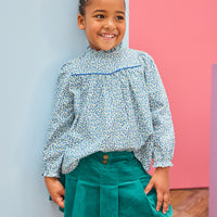 Model can be seen wearing a woven long sleeve top in a blue floral pattern that features a ruffle high neckline and blue trim/pleating detail and button closure along back. Paired back perfectly with a pleated skort in a green velvet to complete the look. 