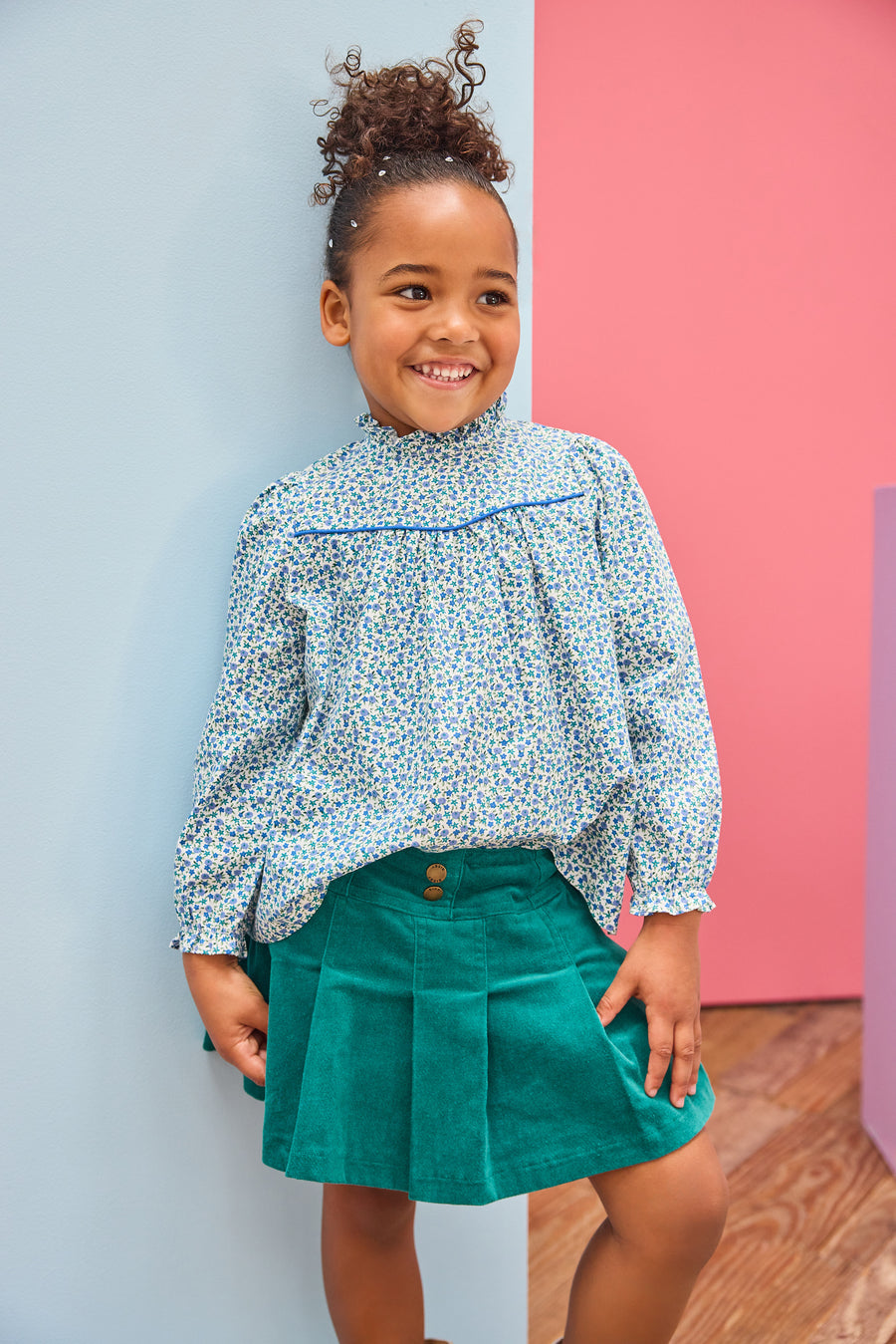 Model can be seen wearing a woven long sleeve top in a blue floral pattern that features a ruffle high neckline and blue trim/pleating detail and button closure along back. Paired back perfectly with a pleated skort in a green velvet to complete the look. 
