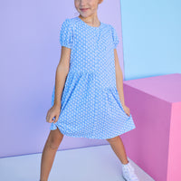 Tween model can be seen wearing a knit dress in a blue/white floral print. Dress features an asymmetrical design, elastic cuffs at sleeves, and button closure along back for an elevated but secure fit. 