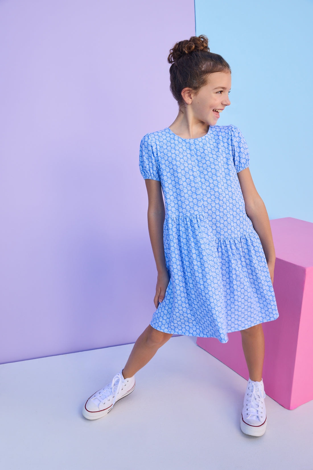 Tween model can be seen wearing a knit dress in a blue/white floral print. Dress features an asymmetrical design, elastic cuffs at sleeves, and button closure along back for an elevated but secure fit. 