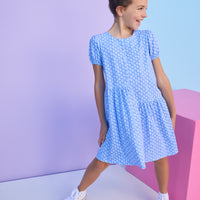 Tween model can be seen wearing a knit dress in a blue/white floral print. Dress features an asymmetrical design, elastic cuffs at sleeves, and button closure along back for an elevated but secure fit. 