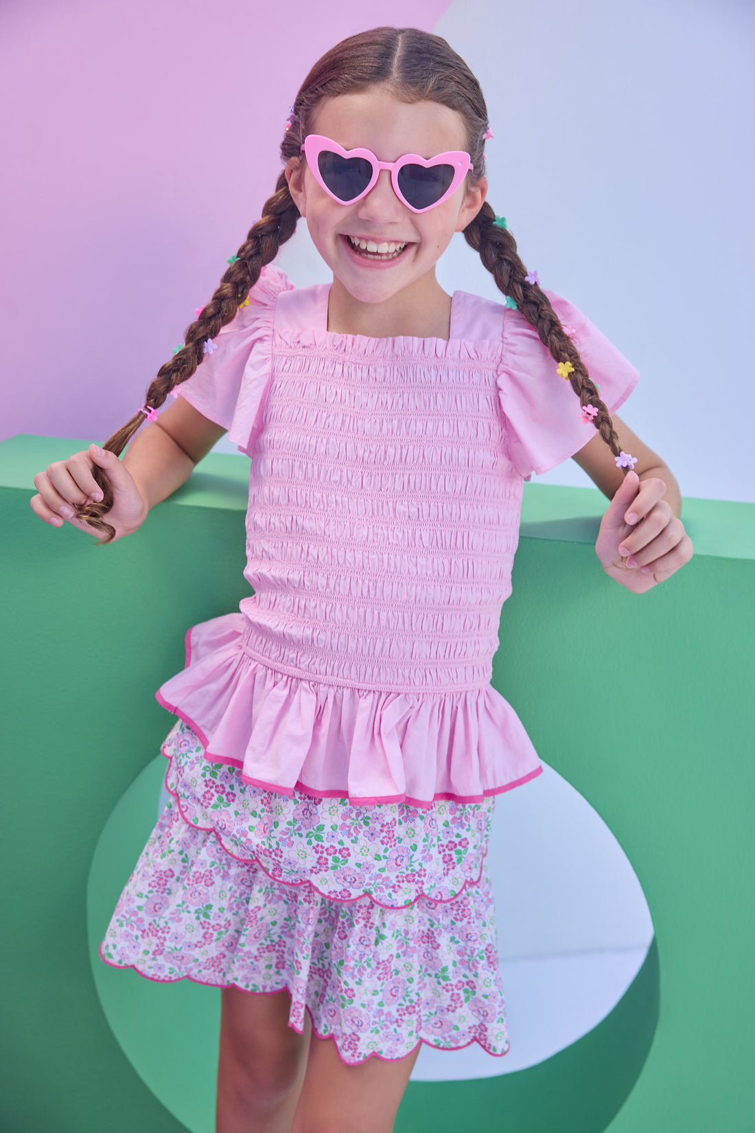 Model can be seen wearing a woven top in a light pink color that features smocking along the whole body of the shirt, angel sleeves, and hot pink trim at bottom to elevate the look. Top pairs back perfectly with tiered skort in a pink floral print to complete the look.