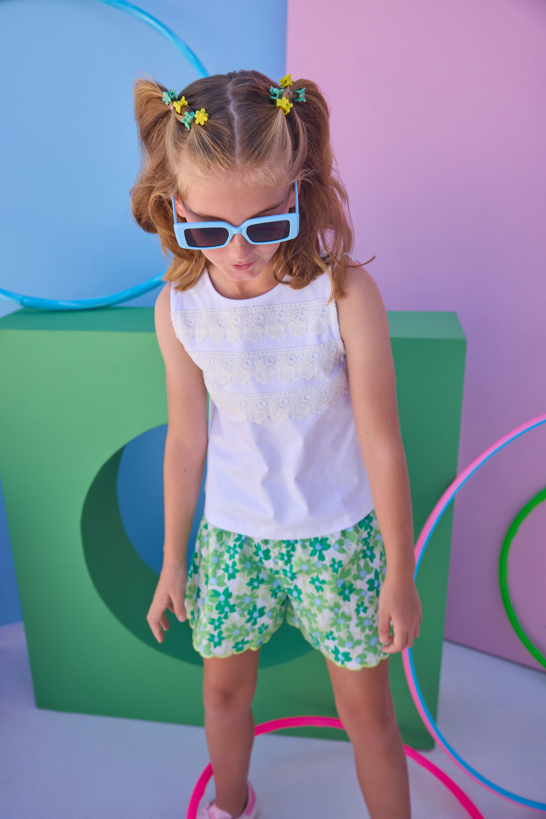 Tween model can be seen wearing shirred shorts in a green/light blue floral print. Shorts feature smocking along waist and scallop edges to elevate the look. Paired back perfectly with a white knit tank top with fun lace features across chest to complete the look. 