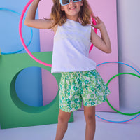 Tween model can be seen wearing shirred shorts in a green/light blue floral print. Shorts feature smocking along waist and scallop edges to elevate the look. Paired back perfectly with a white knit tank top with fun lace features across chest to complete the look. 