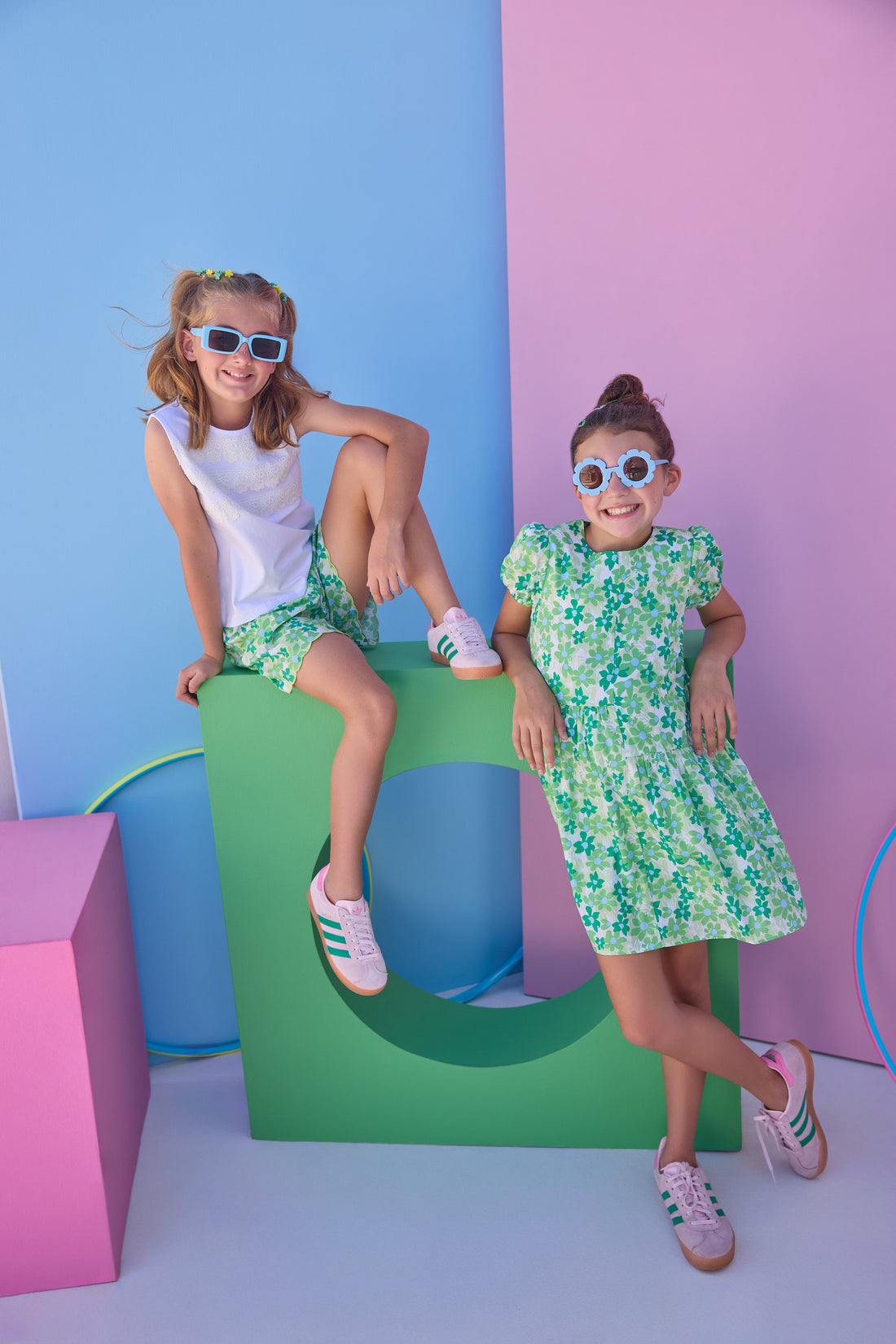 Tween model on left can be seen wearing shirred shorts in a green/light blue floral print. Shorts feature smocking along waist and scallop edges to elevate the look. Paired back perfectly with a white knit tank top with fun lace features across chest to complete the look. Tween model on right can be seen in a knit dress in the same print for a matching moment. 