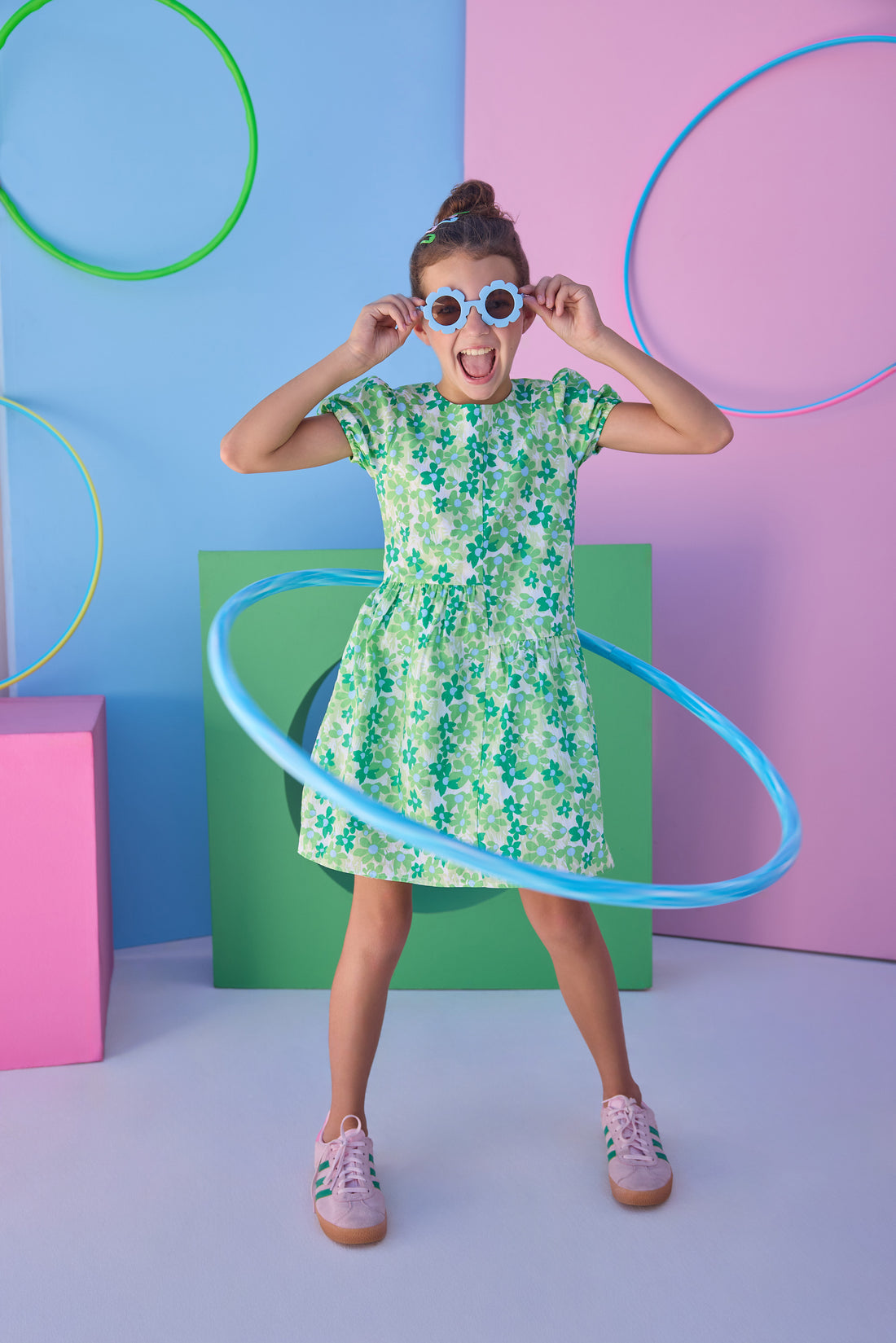 Tween model can be seen wearing a woven dress in a blue and green floral print. Dress features an asymmetrical design, elastic cuffs at sleeves, and button closure along back for an elevated but secure fit. 