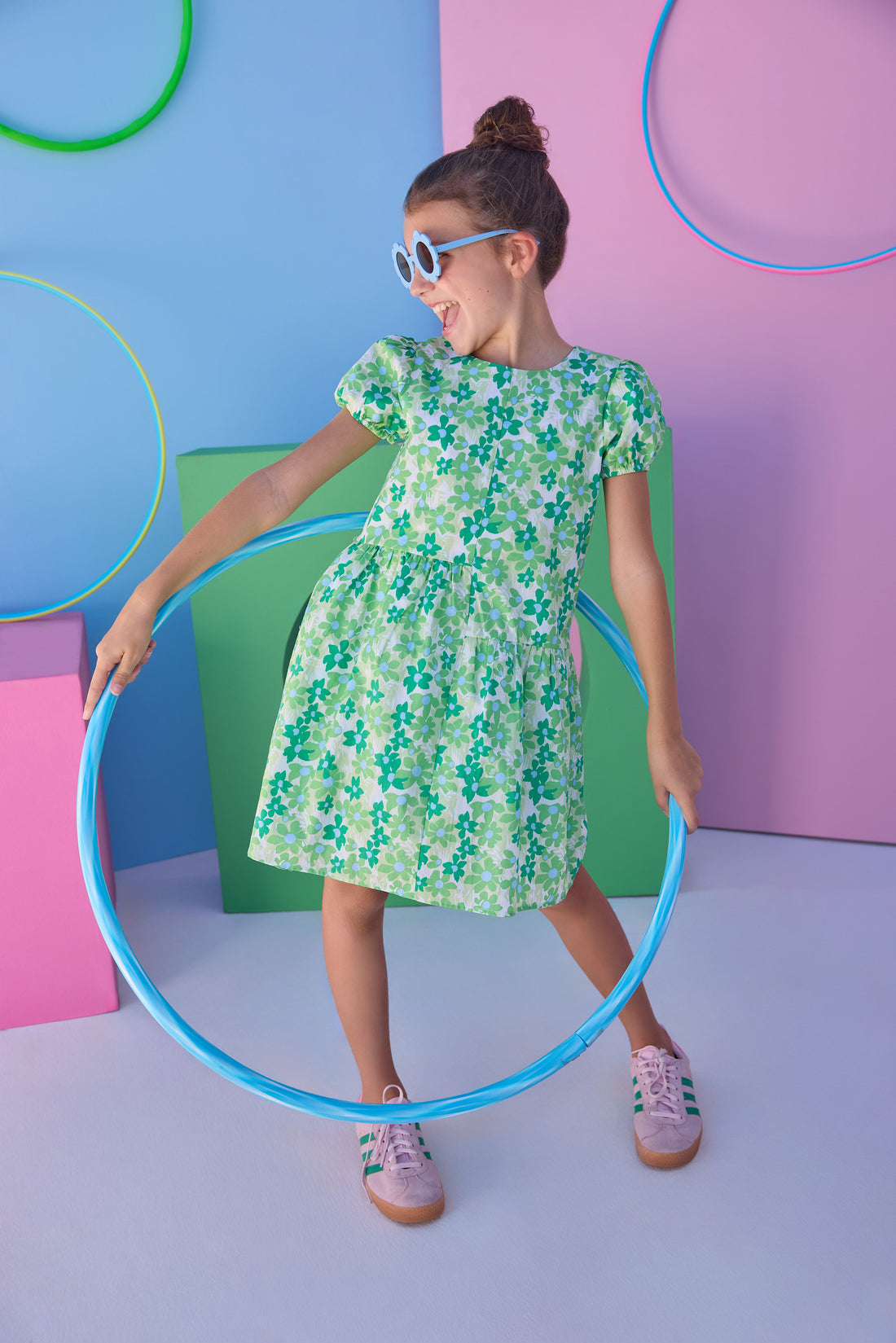 Tween model can be seen wearing a woven dress in a blue and green floral print. Dress features an asymmetrical design, elastic cuffs at sleeves, and button closure along back for an elevated but secure fit. 